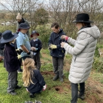 A women in a black hat and long gray coat is showing students how to plant a tree.