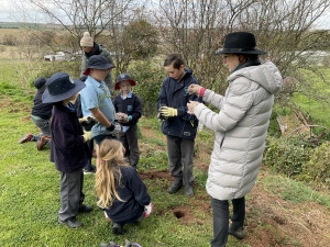 A women in a black hat and long gray coat is showing students how to plant a tree.