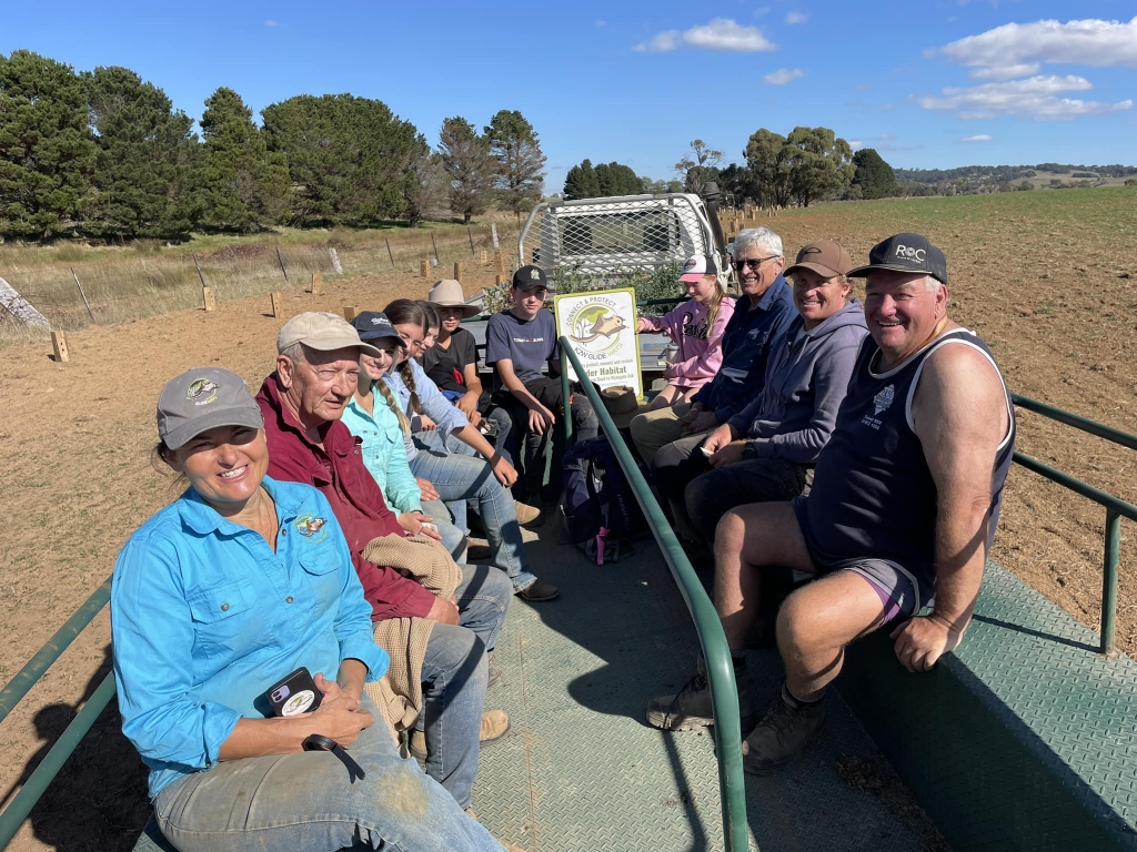 A group of people of all ages on the back of a trailer in a paddock. 